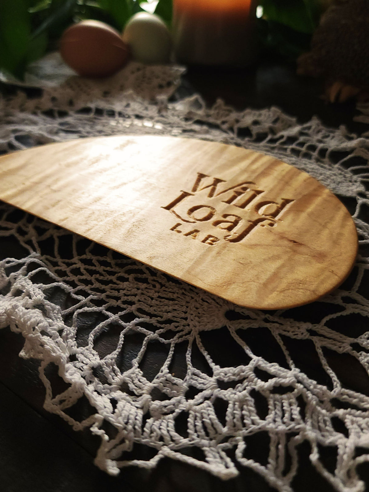 Maple  wooden sourdough bench scraper with 'Wild Loaf Lab" branding on a lace doily with candles and eggs in the background.
