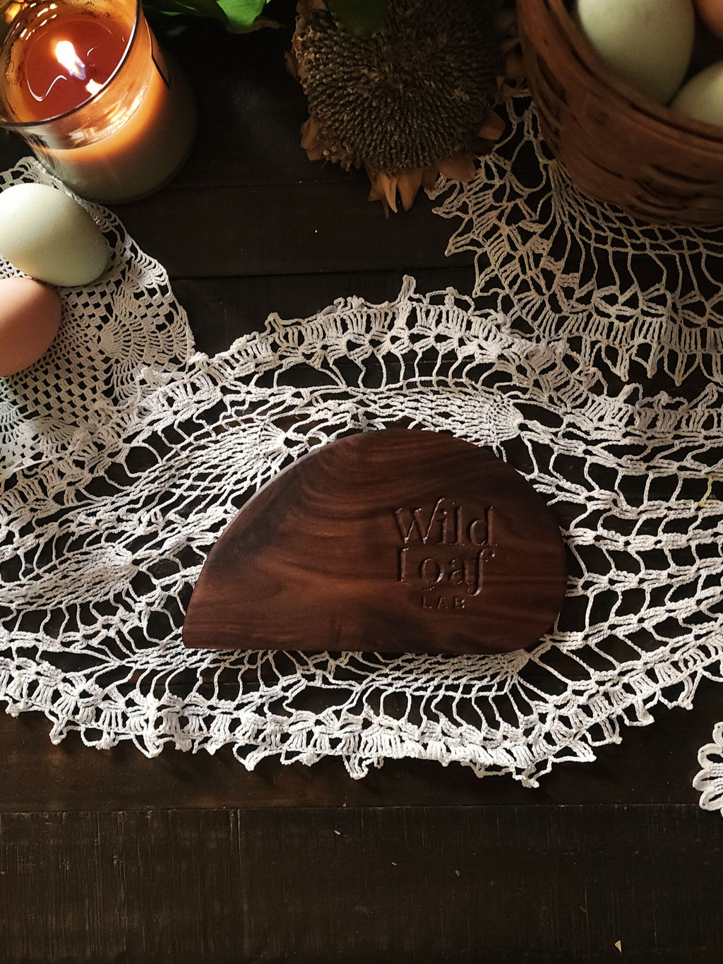 Walnut wooden sourdough bench scraper with 'Wild Loaf Lab" branding on a lace doily with candles and eggs in the background.