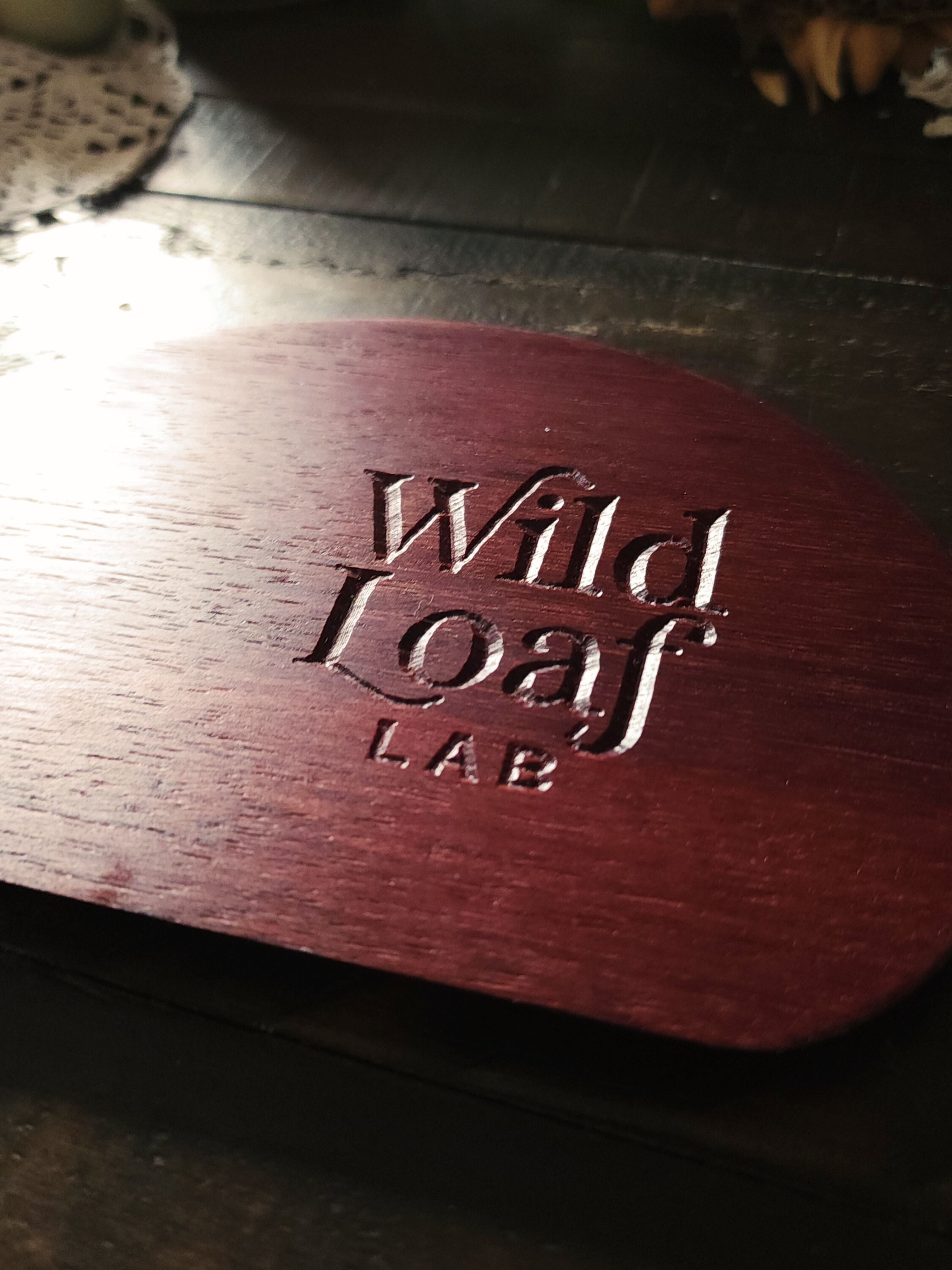 Purpleheart  wooden sourdough bench scraper with 'Wild Loaf Lab" branding on a lace doily with candles and eggs in the background.
