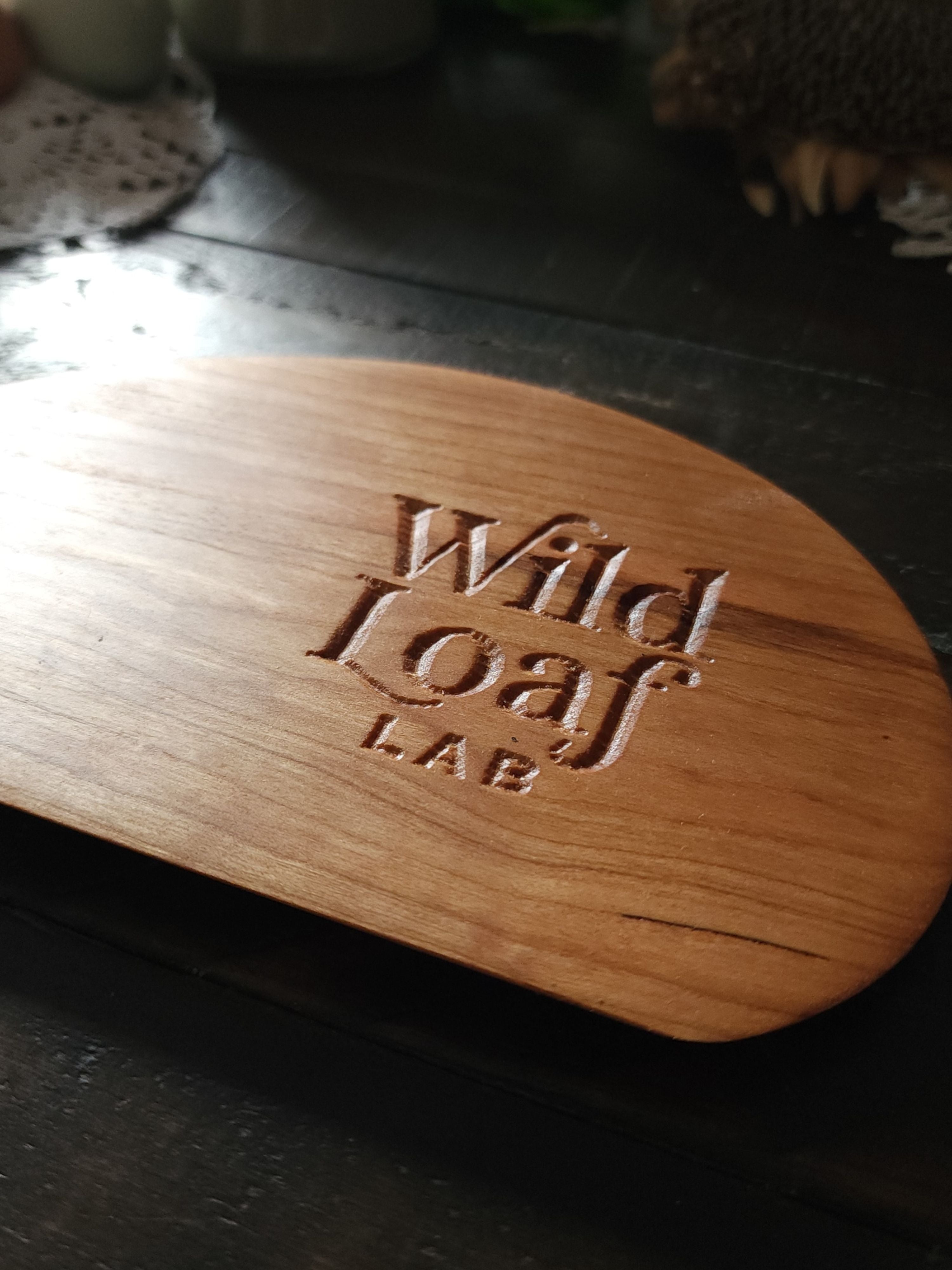 Cherry wooden sourdough bench scraper with 'Wild Loaf Lab" branding on a lace doily with candles and eggs in the background.
