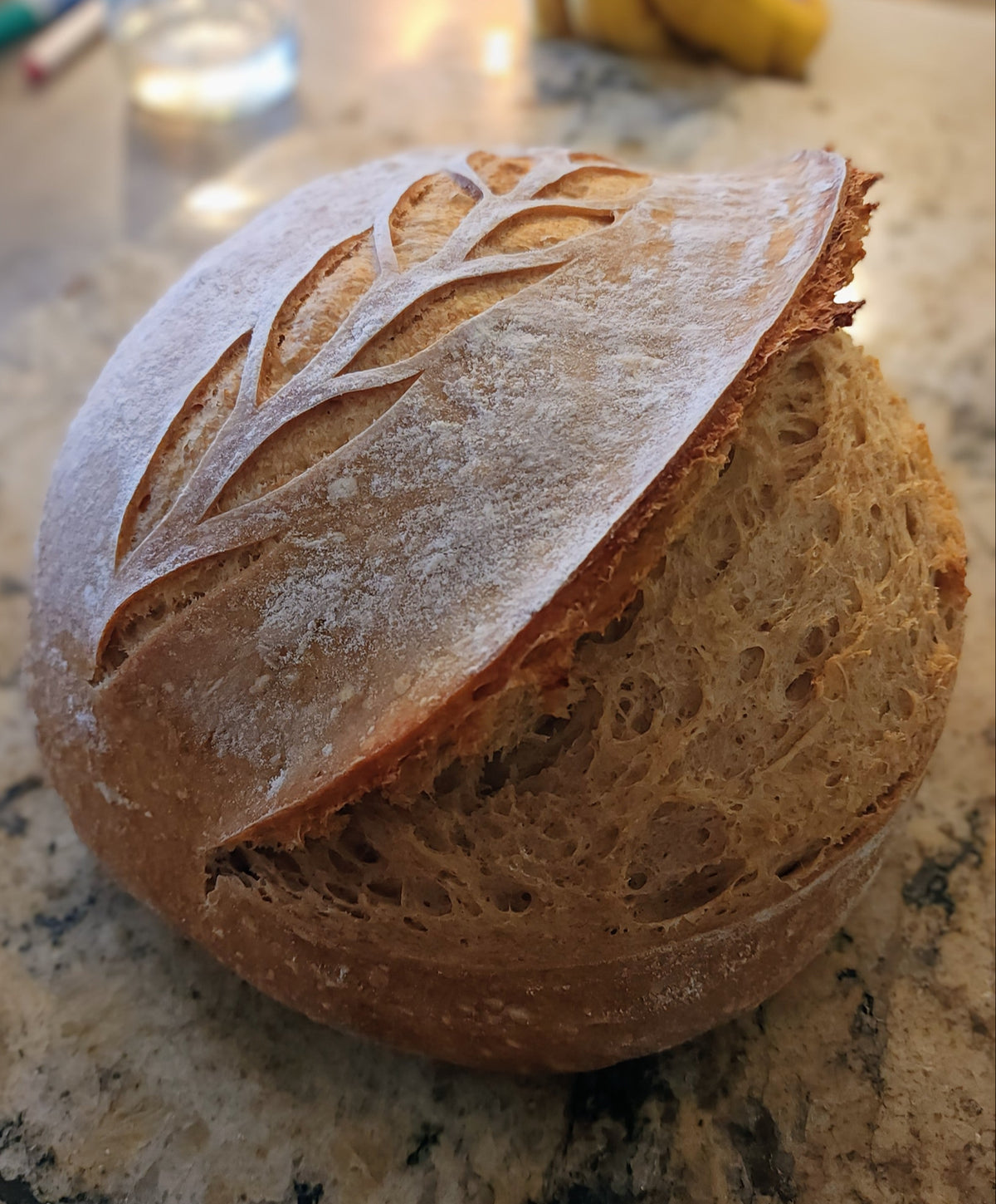 A round artisanal sourdough bread loaf with a scored leaf design on top, sitting on a granite countertop with bananas and a glass jar blurred in the background.